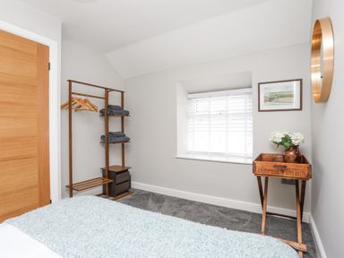 A bedroom corner with a wooden clothes rack holding hangers and folded towels next to a small table with flowers and a window with blinds at Ty Bara in Cemaes Bay