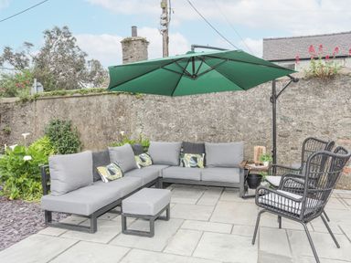 An outdoor seating area with grey cushioned sectional sofas black wicker chairs a green umbrella and a stone wall at Ty Bara in Cemaes Bay