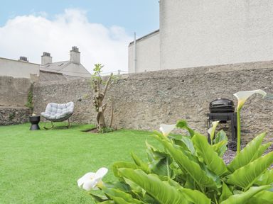 A backyard with green grass a cushioned rocking chair a small black side table a tree a stone wall a barbecue grill and white flowers at Ty Bara in Cemaes Bay