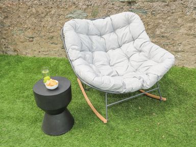 A cushioned rocking chair next to a small black table with a bowl of chips and a glass of lemon water on grass against a stone wall at Ty Bara in Cemaes Bay