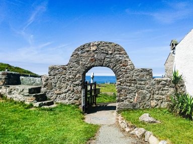 A stone archway with a gate leading to a grassy area with tombstones and the sea in the background at Ty Bara in Cemaes Bay