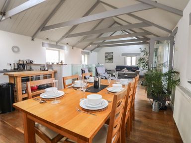 A dining area with a wooden table set for six in an open space with a kitchen island and a sitting area at An Skyber in Perranuthnoe
