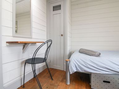 A small bedroom with a single bed a rolled towel on the bed a black metal chair a wall-mounted wooden desk a mirror a door and wooden floor at An Skyber in Perranuthnoe