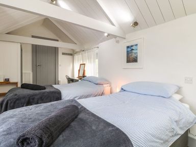 A bedroom with two single beds with striped bedding and rolled towels on top a desk with a mirror and chair near a window with curtains a gray door and a framed picture on the wall at An Skyber in Perranuthnoe