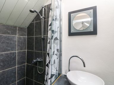 A bathroom with a shower head curtain black tiles a round mirror and a white sink with a faucet at An Skyber in Perranuthnoe