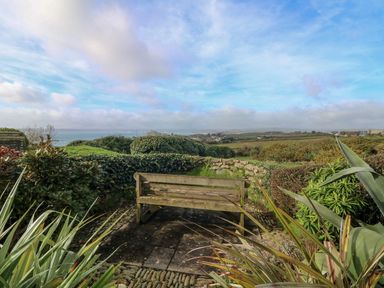 A wooden bench on a stone patio surrounded by plants and overlooking fields and the sea at An Skyber in Perranuthnoe