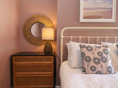 A bedroom corner with a white metal bed frame pillows with flower pattern a wooden nightstand with a lamp and a round mirror on the wall at First Light in Pakefield near Lowestoft