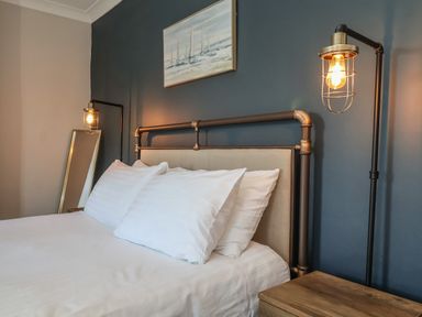 A bed with white pillows and an industrial metal headboard with lit wall lamps and a painting on the wall at First Light in Pakefield near Lowestoft