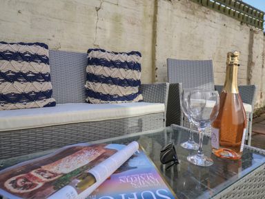 An outdoor seating area with grey wicker furniture cushions a bottle of rose wine two wine glasses a magazine and sunglasses on a glass table at First Light in Pakefield near Lowestoft