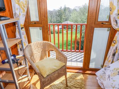 A room with a wicker chair and cushion next to a door leading to a balcony overlooking a garden at Thistle Dubh Doo in Sandyhills