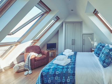 A bedroom with a bed covered with blue patterned bedding a brown armchair a small television and angled windows at Craster Reach in Craster