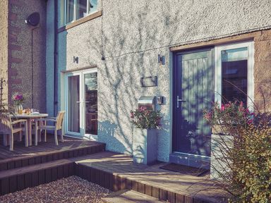 An exterior view of a house with a blue front door a glass sliding door a wooden deck with table and chairs and potted plants at Craster Reach in Craster