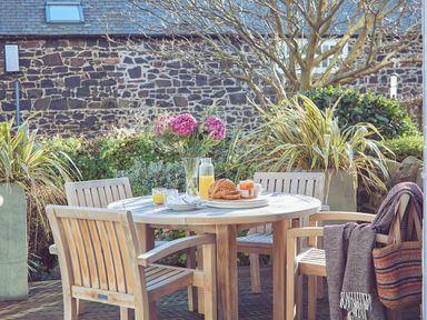 An outdoor wooden table set with orange juice bread and flowers surrounded by wooden chairs at Craster Reach in Craster