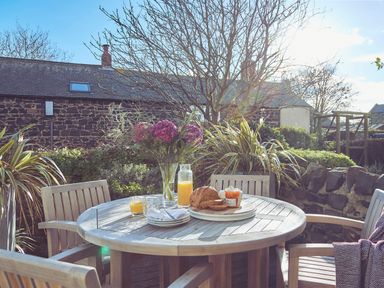 A wooden round table with juice glasses bread jam and flowers on it surrounded by wooden chairs in a garden at Craster Reach in Craster