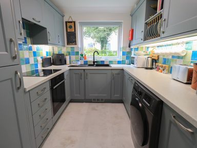 A kitchen with blue cabinets a black oven a washing machine a sink under a window and small appliances on the counters at Little Hallgarth in Keswick