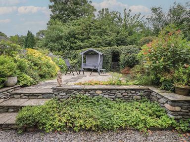 A garden area with stone steps and walls a seated swing two chairs and dense greenery at Little Hallgarth in Keswick