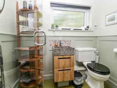 A bathroom with a wooden sink cabinet black toilet seat shelving unit and window at Little Hallgarth in Keswick