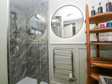 A bathroom with a glass shower enclosure grey tiled walls a round mirror a chrome towel rack and wooden shelves with toiletries at Little Hallgarth in Keswick