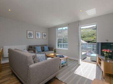 A living room with two sofas a coffee table and a TV on a stand with open door to a balcony showing a hillside outside at Rebecca in Mullion Cove near Mullion