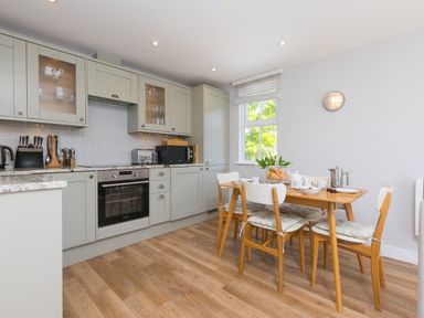 A kitchen with gray cabinets wooden dining table and chairs set for breakfast near a window with white blinds at Rebecca in Mullion Cove near Mullion