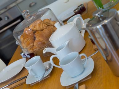 A wooden table set with white teacups saucers a teapot a glass dome covering croissants and small jars of jam at Rebecca Mullion Cove near Mullion