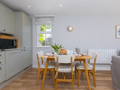 A dining area with a wooden table set for breakfast and a kitchen with grey cabinets and a microwave at Rebecca in Mullion Cove near Mullion