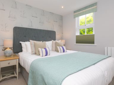 A bedroom with a double bed a green blanket and pillows two bedside tables with lamps and a window with a green blind at Rebecca Mullion Cove near Mullion