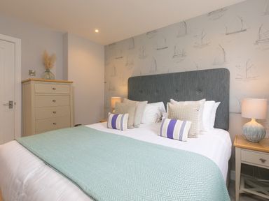 A bedroom with a bed featuring a blue headboard and patterned pillows with a chest of drawers and bedside tables with lamps at Rebecca in Mullion Cove near Mullion