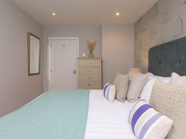 A bedroom with a bed featuring multiple pillows a dresser with a vase and pampas grass a wall mirror and sailing boat wallpaper at Rebecca in Mullion Cove near Mullion