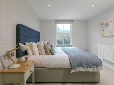 A bedroom with a bed multiple pillows a side table with a lamp and flower and a window at Rebecca in Mullion Cove near Mullion