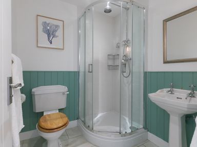 A bathroom with a corner glass shower, toilet with wooden seat, sink with mirror and framed artwork at Rebecca in Mullion Cove near Mullion