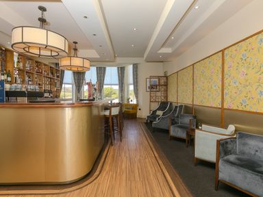A bar area with stools and shelves of bottles beside a seating area with chairs and floral wallpaper at Rebecca in Mullion Cove near Mullion