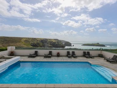 An outdoor swimming pool with lounge chairs and potted plants overlooking a rocky coastline and the sea at Rebecca in Mullion Cove near Mullion