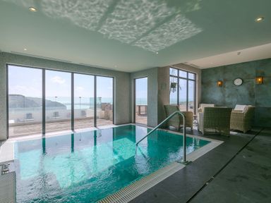 An indoor swimming pool with glass windows showing an ocean view and wicker chairs in a seating area at Rebecca Mullion Cove near Mullion