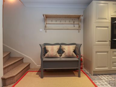 A gray wooden bench with two pillows below a wall-mounted coat rack next to carpeted stairs and kitchen cabinets at Millpond Bothy in Woolhope