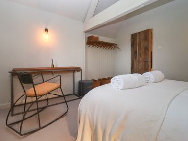A bedroom with a double bed with rolled towels a wooden desk with a rocking chair a wooden door and wall-mounted coat rack at Millpond Bothy in Woolhope