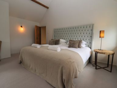 A bedroom with a large bed with beige blanket and rolled white towels a wooden door a wall light and a wooden side table with a lamp at Millpond Bothy in Woolhope