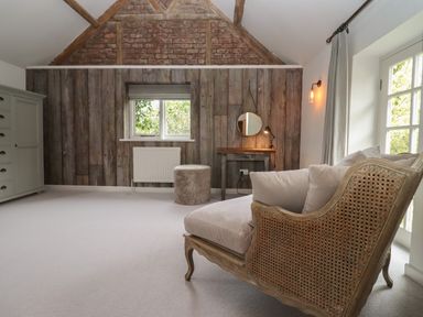 A room with a wicker armchair cushions a small table with a round mirror a pouf a cupboard and a window at Millpond Bothy in Woolhope