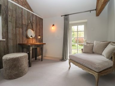 A room with a wooden table, round mirror, upholstered stool, cushioned wooden chair, and a door with a curtain at Millpond Bothy in Woolhope