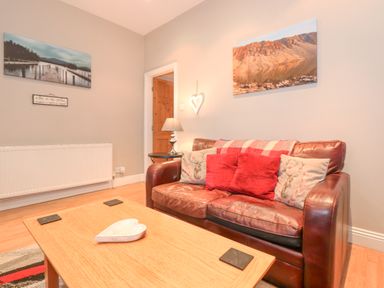 A living room with a brown leather sofa with cushions a wooden coffee table with a heart-shaped dish a lamp on a side table and landscape photos on the walls at Myrtle Cottage in Windermere
