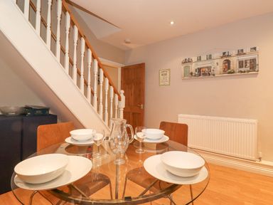 A dining area with a glass table set for four under a staircase with wooden chairs and wall art at Myrtle Cottage in Windermere