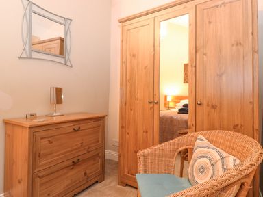A room with a wooden chest of drawers mirror wardrobe and wicker chair at Myrtle Cottage in Windermere