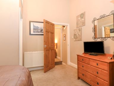 A bedroom with a wooden dresser holding a television a mirror above two paintings on the wall a partially open wooden door and a framed photo above a radiator at Myrtle Cottage in Windermere