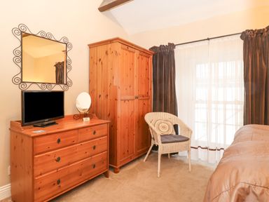 A bedroom with a wooden dresser holding a television and mirror a wooden wardrobe a wicker chair with a cushion and a bed with brown bedding and sheer curtains at Myrtle Cottage in Windermere