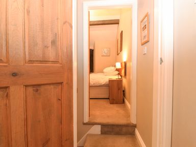 View of a bedroom with a bed lamp and wooden nightstand through a hallway with carpeted steps and wooden door at Myrtle Cottage in Windermere