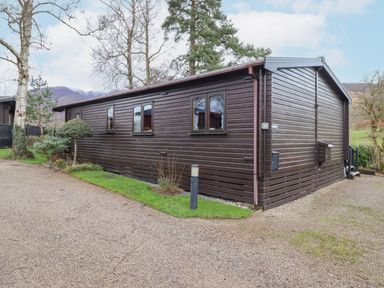 A dark brown wooden cabin with windows and a small garden area on a gravel path at Hayeswater Lodge in Burnside Park near Keswick