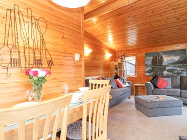 A living and dining area with wooden walls and ceiling a dining table with chairs a vase of flowers two gray sofas an ottoman and a large landscape painting at Hayeswater Lodge Burnside Park near Keswick
