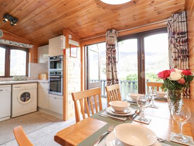A dining table set with plates bowls glasses and cutlery next to large windows and a kitchen with appliances at Hayeswater Lodge Burnside Park near Keswick