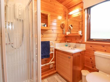 A bathroom with a shower, sink with wooden cabinet, toilet, window, and wooden walls at Hayeswater Lodge in Burnside Park near Keswick