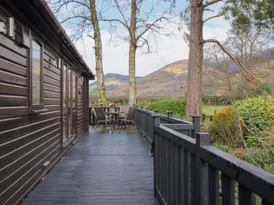 An outdoor wooden deck with a table and chairs next to a dark wood cabin with mountains and trees in the background at Hayeswater Lodge Burnside Park near Keswick